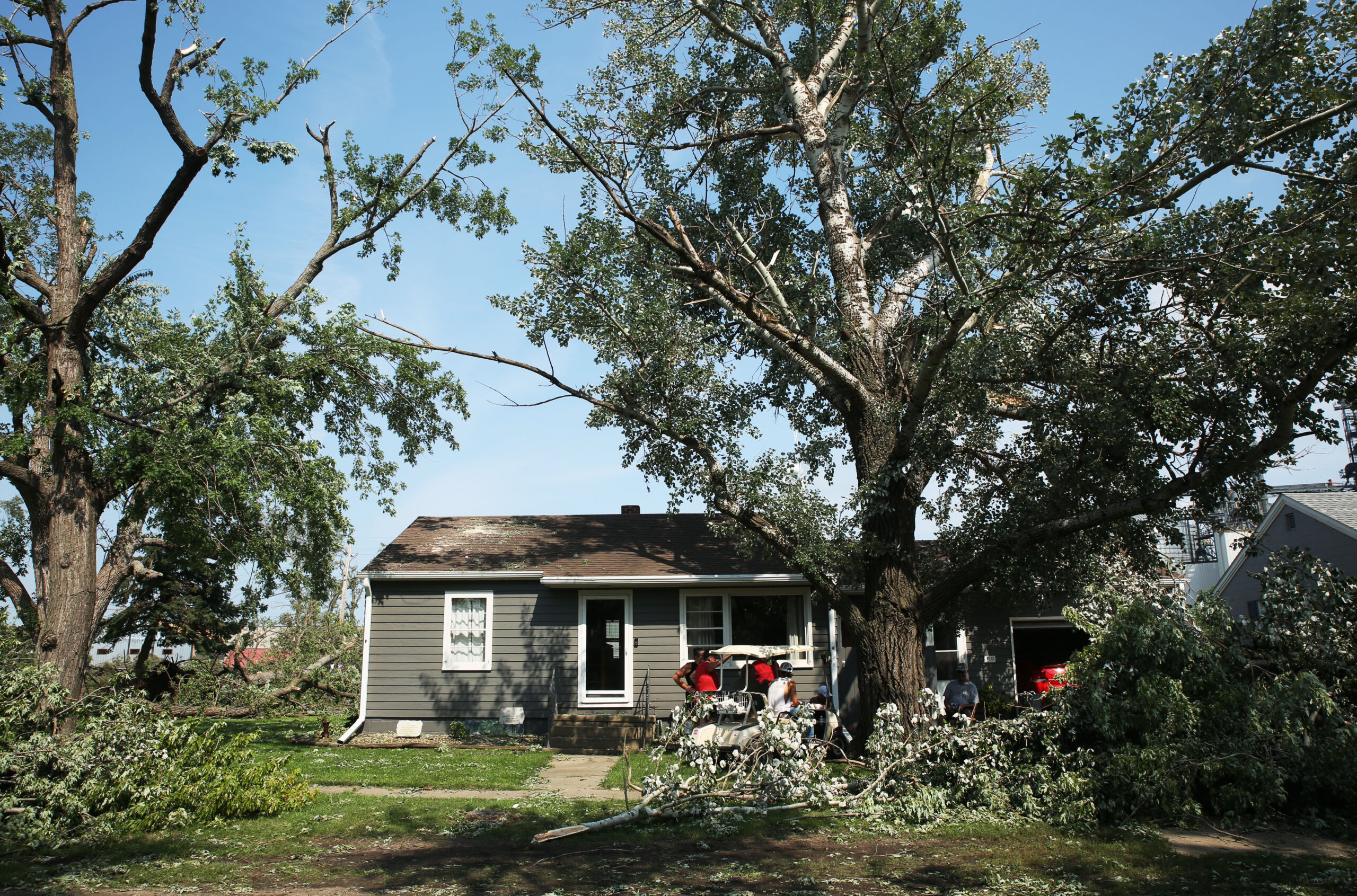 Hudson residents gather under a patch of shade Tuesday, July 29, 2025 as they take a break from cleaning up debris after severe weather moved through the area Monday evening damaging trees, homes and other structures. (Makenzie Huber/South Dakota Searchlight)