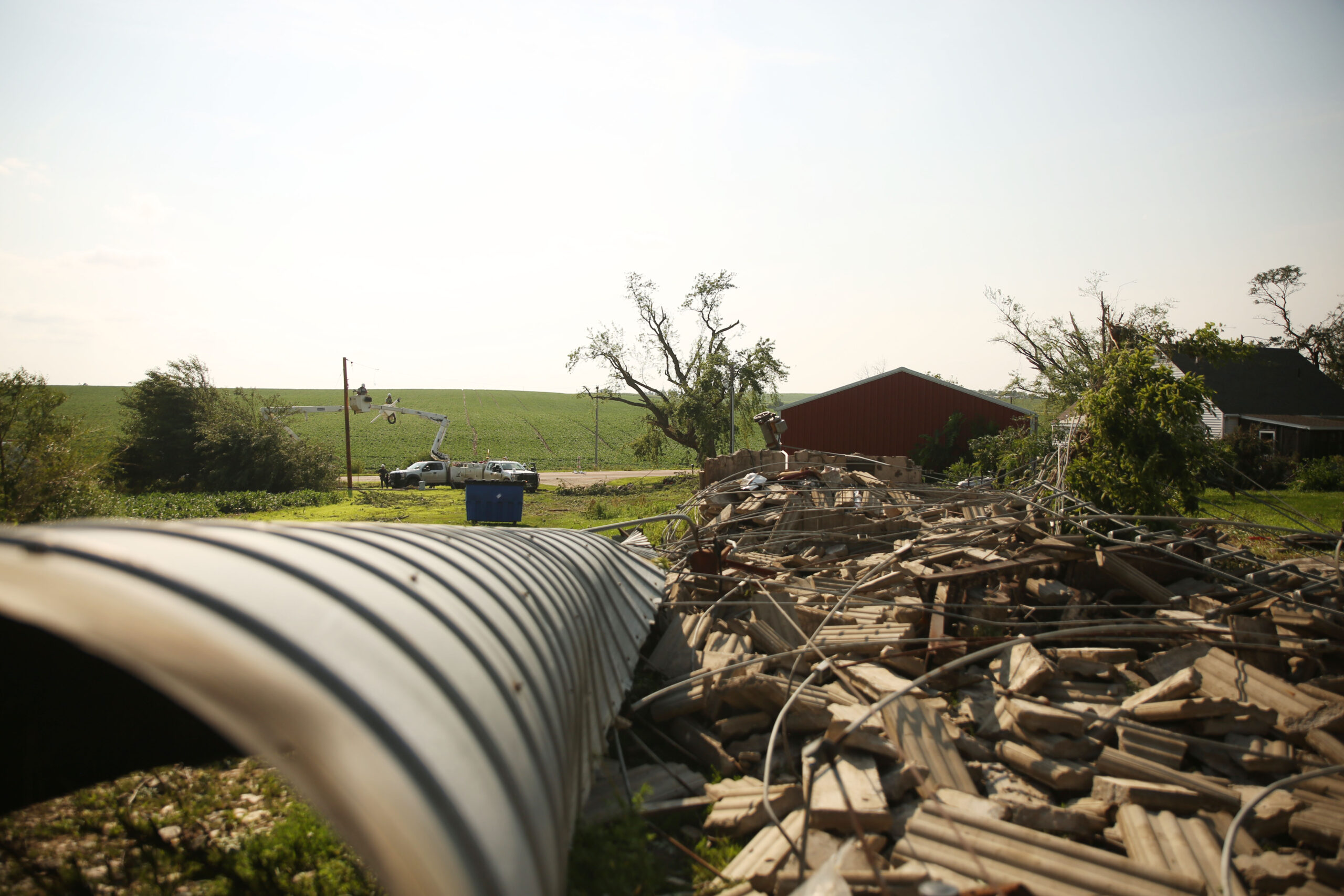 Workers repair powerlines just outside of Hudson as Amanda Lounsbery surveys damage on her property on Tuesday, July 29, 2025. Wind knocked over Lounsbery's silo, shattering concrete and splitting the structure. Severe weather moved through the area Monday evening. The system damaged trees, homes and other structures. (Makenzie Huber/South Dakota Searchlight)