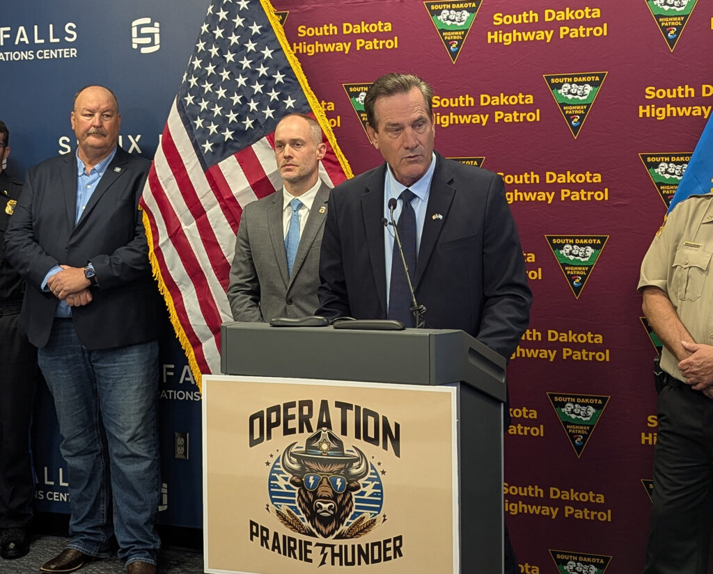 Gov. Larry Rhoden speaks to reporters from a podium on July 28, 2025, at the Public Safety Administration Building in Sioux Falls. Also visible are, from left, Dan Satterlee, director of the state Division of Criminal Investigation, and Sam Olson, Minneapolis field office director for U.S. Immigration and Customs Enforcement. (John Hult/South Dakota Searchlight)