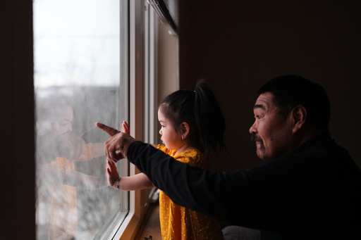 AvaRae Carl looks out of the window of her family's temporary housing in Bethel, Alaska, with her grandfather Joe Paul as he points in the direction of Kipnuk village, Tuesday, Oct. 28, 2025, after they were displaced from their home due to Typhoon Halong that hit earlier in the month. (AP Photo/Lindsey Wasson)